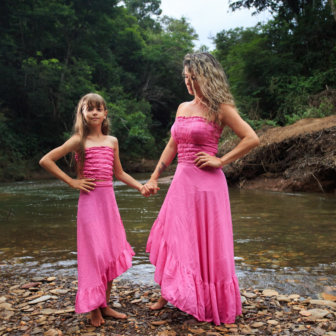 Mãe e filha de mãos dadas na beira do rio, com vestidos cor-de-rosa, cuidado em família com cosméticos naturais