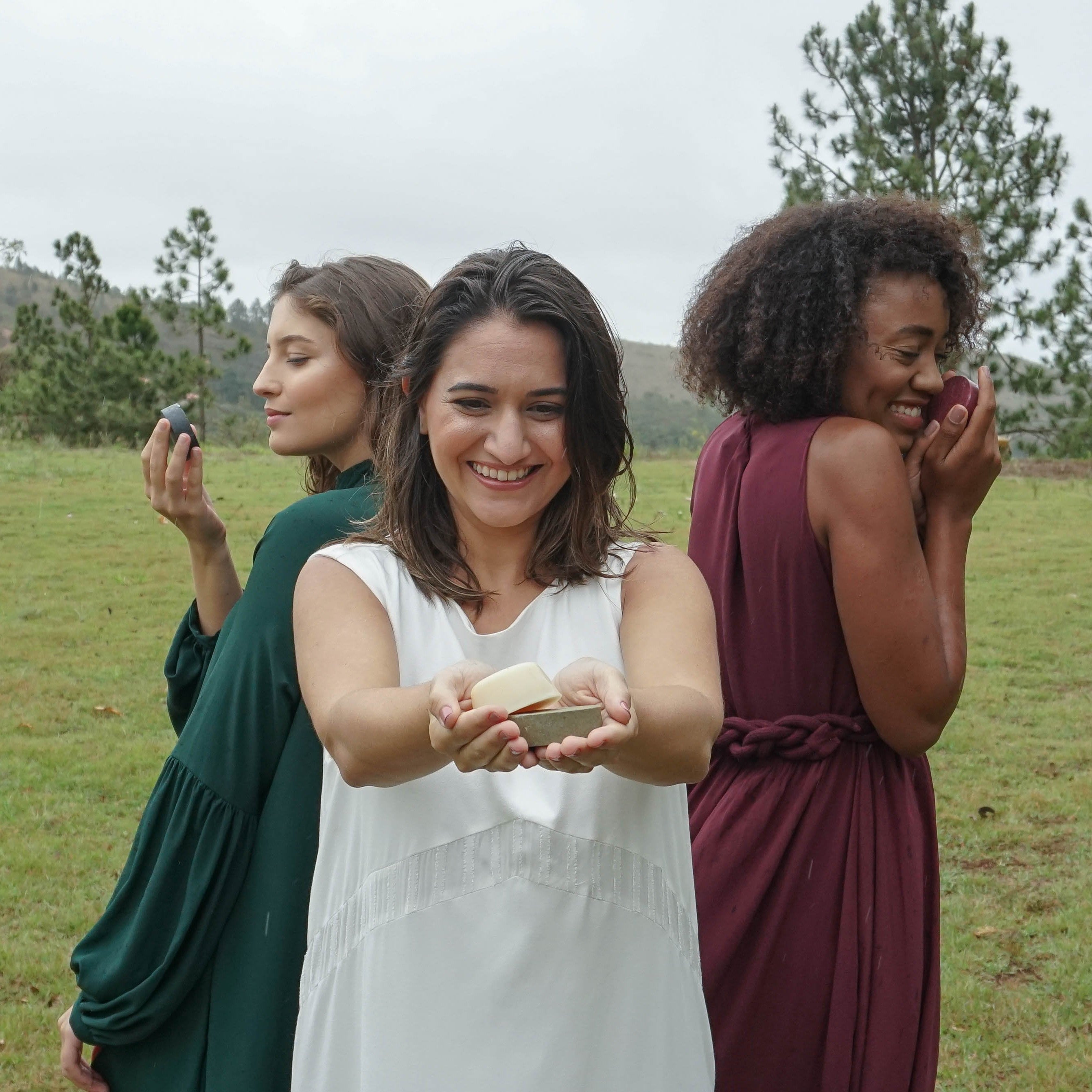 Três mulheres com cabelos e estilos diferentes segurando barras de shampoo OR Natural ao ar livre